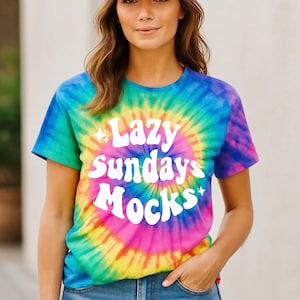 May include: A tie-dye t-shirt with the text "Lazy Sundays Mocks" in white, paired with denim shorts and a pink baseball cap. The shirt features a spiral design with vibrant colors including blue, green, yellow, and pink.