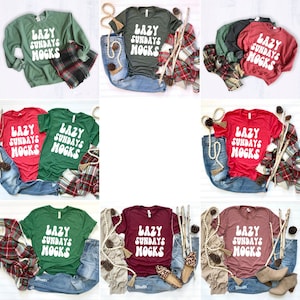 May include: Nine different t-shirts featuring the text "Lazy Sundays Mocs" in a retro font. The shirts are in a variety of colours, including red, green, maroon, and pink. The shirts are laid out on a white surface with various props, such as jeans, scarves, and pinecones.