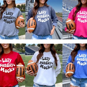 K&ouml;nnte beinhalten: Eine Auswahl an Sweatshirts mit Rundhalsausschnitt in verschiedenen Farben, darunter Grau, Marineblau, Pink, Rot, Wei&szlig; und Blau. Jedes Sweatshirt zeigt den Text "Lazy Sundays Mocks" in einer Retro-Schriftart. Ein brauner Football wird vor jedem Sweatshirt gehalten.