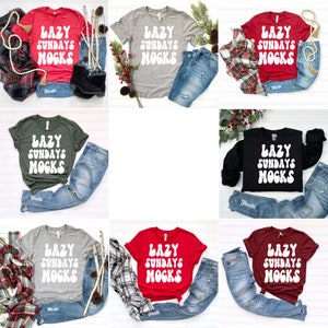 May include: Nine different coloured t-shirts with the text "LAZY SUNDAYS MOCKS" in white, retro-style lettering. The shirts are displayed on a white surface with jeans and other accessories.