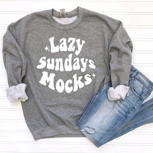May include: A gray sweatshirt with the text "Lazy Sundays Mocks" in white, paired with blue denim jeans. The sweatshirt's sleeves are cuffed, revealing a lighter gray interior. The jeans have distressed details.