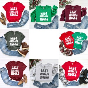 May include: A collection of nine t-shirts featuring the text "Lazy Sundays MOGKS" in a retro font. The shirts are in a variety of colours, including red, green, maroon, and grey. The shirts are displayed on a white surface with jeans, boots, and other accessories.
