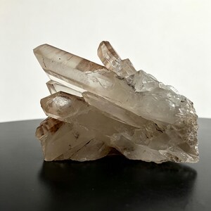 May include: A cluster of translucent, smoky quartz crystals featuring sharp, elongated points and natural facets. The mineral formation displays varying shades of pale brown and clear white, with a jagged, crystalline texture throughout the structure.
