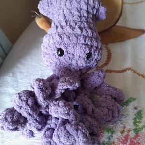 Puede incluir: Un juguete de peluche de pulpo de ganchillo hecho a mano, de color lavanda. El pulpo tiene un aspecto texturizado y esponjoso con ojos de seguridad negros y múltiples tentáculos. El peluche está sobre una superficie blanca con un estampado floral.