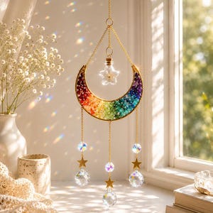 May include: A rainbow-colored crescent moon suncatcher with gold-colored metal accents. The moon is filled with colorful stones and hangs from a gold chain. Below the moon are hanging crystals and gold stars. A small star-shaped glass bottle hangs from the top.