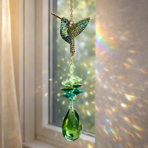 May include: A decorative hummingbird suncatcher with a gold chain. The hummingbird is embellished with green and blue crystals. Below the hummingbird are green crystal beads and a large green teardrop-shaped crystal. The suncatcher hangs in front of a window.