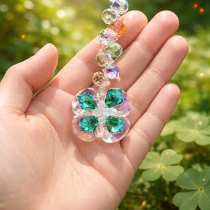 May include: A clear crystal suncatcher with a four-leaf clover design, featuring teal and clear faceted crystals. The suncatcher is suspended from a string of smaller, colorful crystals. The background is a blurred view of green foliage.