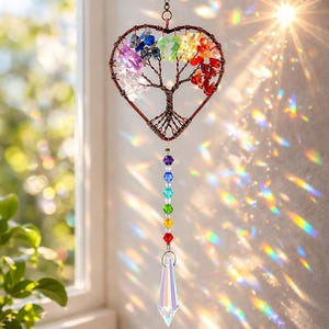 Puede incluir: Un atrapasoles en forma de corazón con un diseño de árbol, elaborado con alambre de cobre y piedras preciosas de colores. Un arcoíris de cristales cuelga debajo, culminando en un colgante de cristal claro y facetado. El atrapasoles está suspendido frente a una ventana.