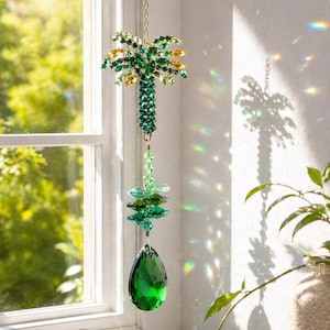 Puede incluir: Un atrapasoles colgante con forma de palmera, con cristales verdes y amarillos. El atrapasoles está suspendido por una cadena dorada y presenta un gran cristal verde en forma de lágrima en la parte inferior. El atrapasoles está colgado frente a una ventana.
