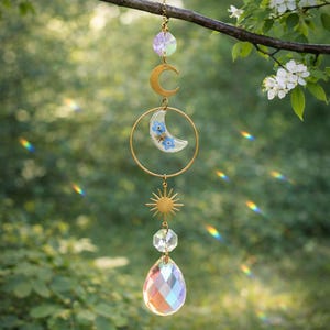 May include: A celestial-themed suncatcher with a faceted crystal teardrop, a sunburst, a gold-coloured crescent moon, and a clear moon-shaped pendant with blue flowers. The suncatcher is suspended from a branch.