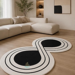 Op de afbeelding: Een modern, abstract tapijt in een achtvormig ontwerp. Het tapijt heeft een crèmekleurige achtergrond met zwarte en witte gebogen lijnen en zwarte cirkelvormige centra. Het tapijt ligt op een lichte houten vloer in een woonkamer.