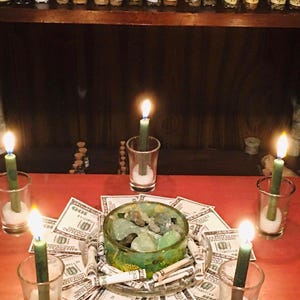 May include: A collection of small glass jars with cork stoppers arranged on a shelf above a red surface. Five green candles in clear glass votives surround a central glass bowl filled with green stones and surrounded by one hundred dollar bills.