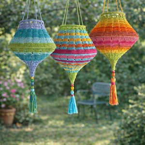 Puede incluir: Tres faroles de ganchillo hechos a mano y coloridos, colgados de una rama de árbol. Cada farol presenta una combinación de colores única, incluyendo azul, verde, naranja y rosa, con una borla y cuentas en la parte inferior. Los faroles están en un jardín.