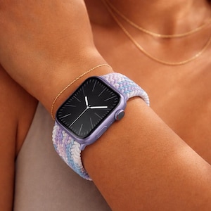 Könnte beinhalten: Eine lavendelfarbene Smartwatch mit schwarzem Bildschirm und weißen Zeigern wird am Handgelenk getragen. Das Uhrenarmband ist eine Pastellmischung aus Blau, Weiß und Lavendel. Ein goldenes Armband und eine Halskette sind ebenfalls sichtbar.