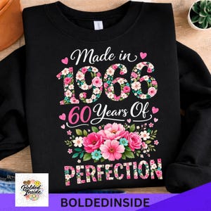 Może przedstawiać: Czarna bluza z napisem "Made in 1966 60 Years Of Perfection". Cyfry zdobione wzorami kwiatowymi w kolorze różowym, białym i zielonym. Różowe serca i kwiaty podkreślają design. Nazwa marki "Bolded Inside" znajduje się na dole.