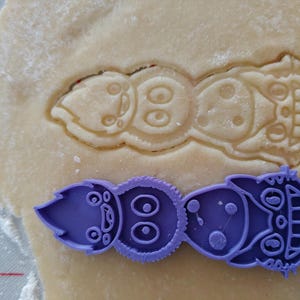 May include: A purple plastic cookie cutter in the shape of a whimsical creature, next to a cutout of the same design in dough. The cutter has a detailed design with eyes, dots, and a smiling face. The dough is light brown and dusted with flour.