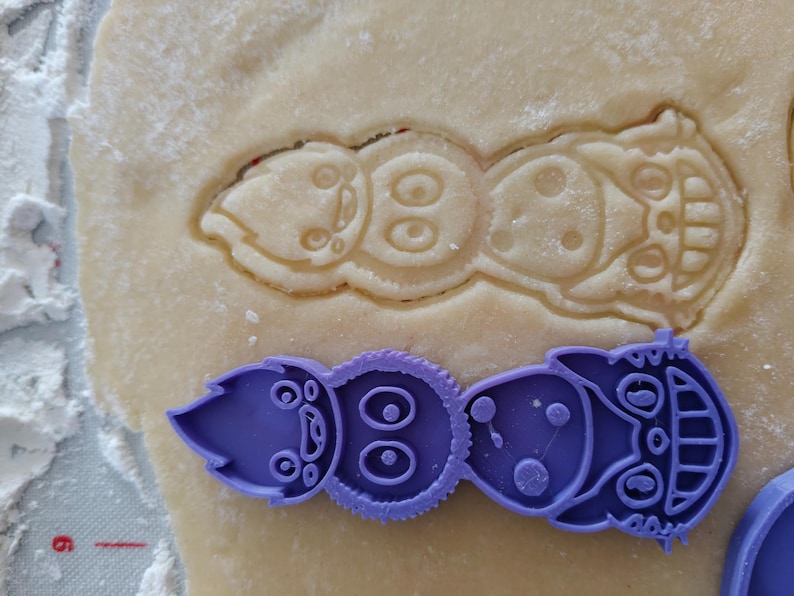 May include: A purple plastic cookie cutter in the shape of a whimsical creature, next to a cutout of the same design in dough. The cutter has a detailed design with eyes, dots, and a smiling face. The dough is light brown and dusted with flour.