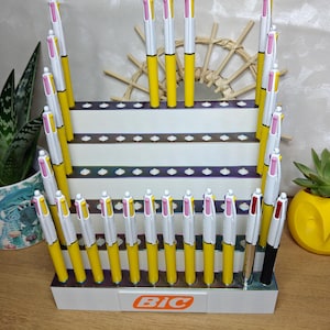 May include: A white tiered retail display stand featuring the Bic logo in orange, holding multiple multi-color ink pens with yellow barrels, white clips, and pink, yellow, and white top buttons, arranged in rows on a wooden surface.