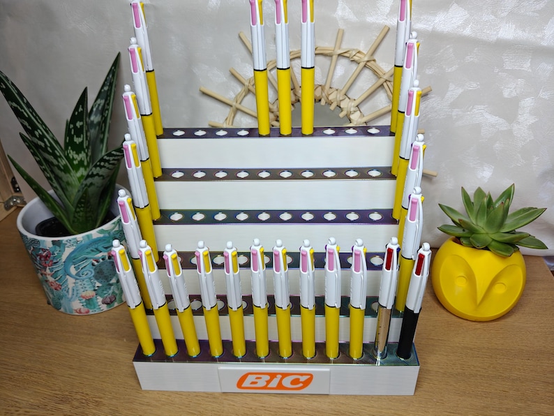 May include: A white tiered retail display stand featuring the Bic logo in orange, holding multiple multi-color ink pens with yellow barrels, white clips, and pink, yellow, and white top buttons, arranged in rows on a wooden surface.