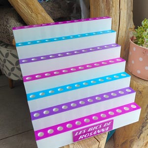 May include: A white tiered display stand with five levels, each featuring a row of circular holes for holding items. The edges of the tiers alternate between bright pink and blue. A pink rectangular plaque reads LES BICS DE ROSANNA.