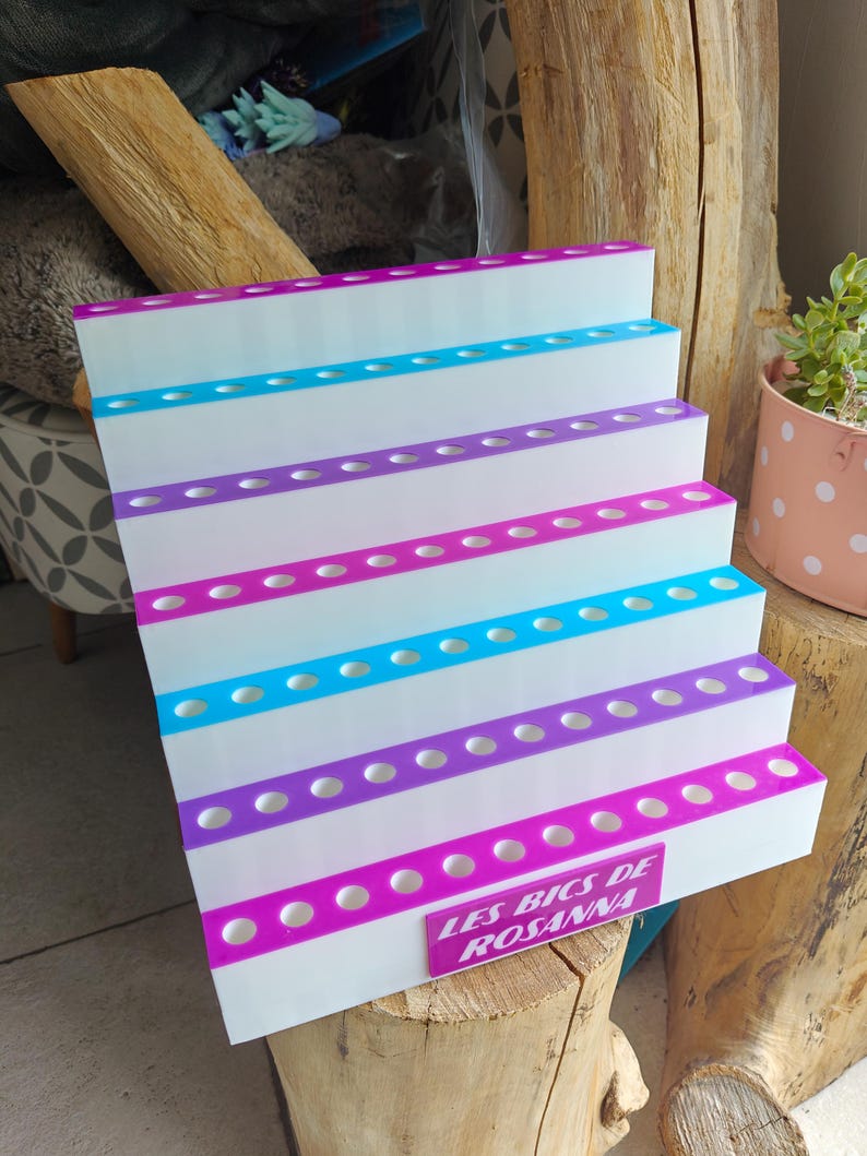 May include: A white tiered display stand with five levels, each featuring a row of circular holes for holding items. The edges of the tiers alternate between bright pink and blue. A pink rectangular plaque reads LES BICS DE ROSANNA.