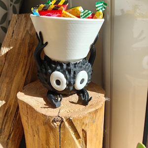 May include: A whimsical black and white candy bowl holder shaped like a cartoon character, holding a white bowl filled with colorful wrapped candies. The holder has large white eyes and is perched on a wooden stump.