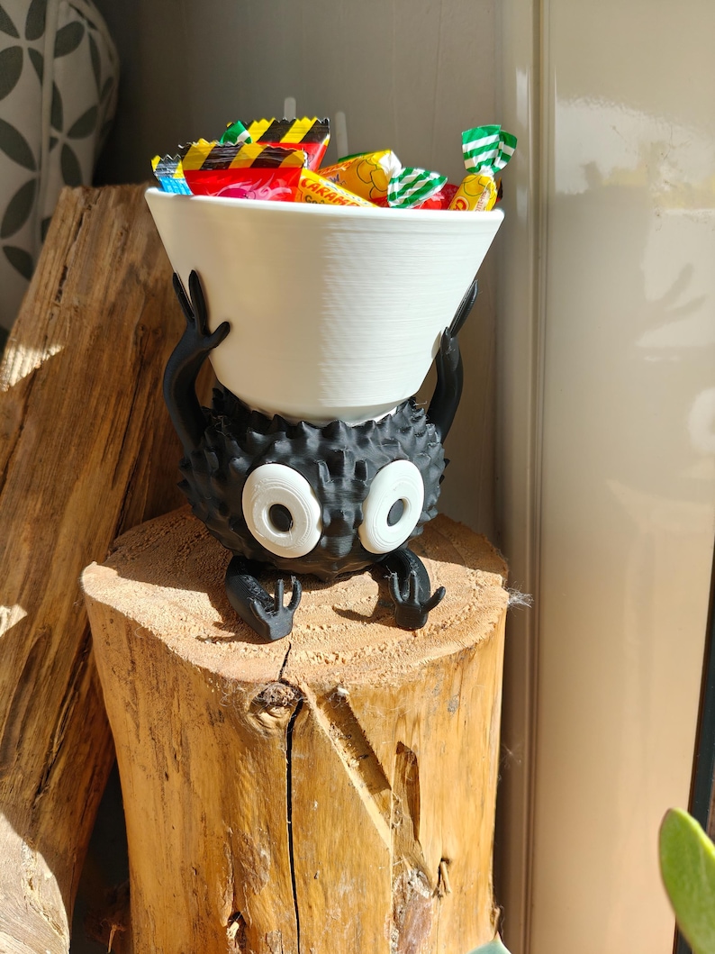 May include: A whimsical black and white candy bowl holder shaped like a cartoon character, holding a white bowl filled with colorful wrapped candies. The holder has large white eyes and is perched on a wooden stump.