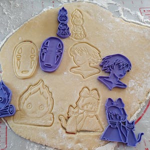 May include: A sheet of dough with various purple cookie cutters in the shapes of characters from a popular animated film. The cut dough shows the impressions of the characters. The scene is on a white surface.