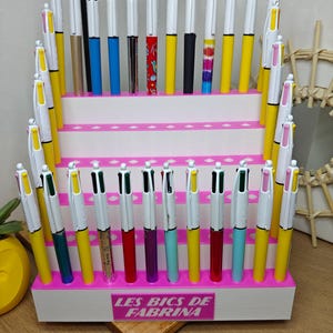 Puede incluir: Una exhibición de bolígrafos coloridos con barriles blancos, detalles amarillos y de colores, y tinta negra. Los bolígrafos están dispuestos en un expositor rosa y blanco escalonado. El texto "LES BICS DE FABRINA" es visible en la parte delantera.