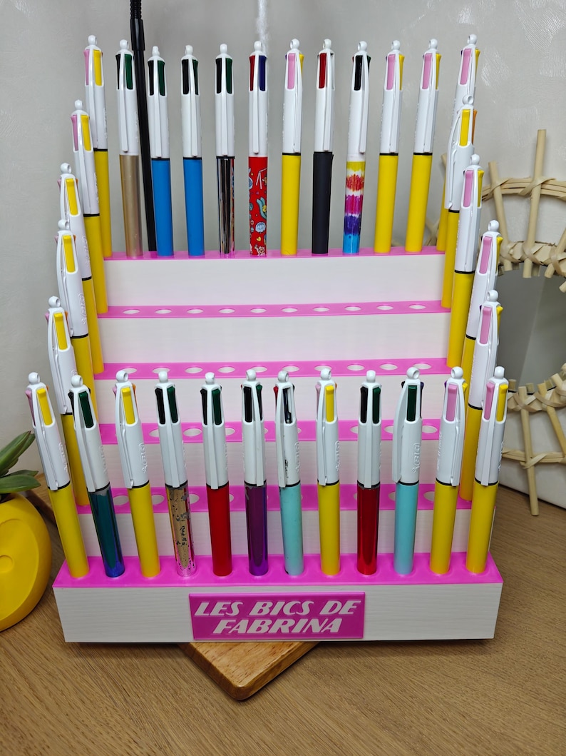 May include: A white tiered display stand with pink accents holding numerous multi-color ink pens. The base features a pink rectangular sign with the white text LES BICS DE FABRINA. The pens have white caps and various colored barrels including yellow, blue, red, and patterned designs.