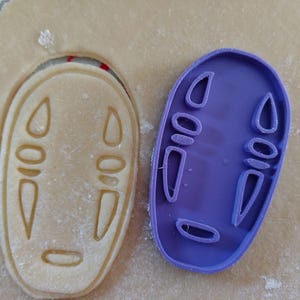 May include: A purple plastic cookie cutter in the shape of a character's face from the animated film, Spirited Away, next to a cookie cut out of the same shape. The cookie is light brown.