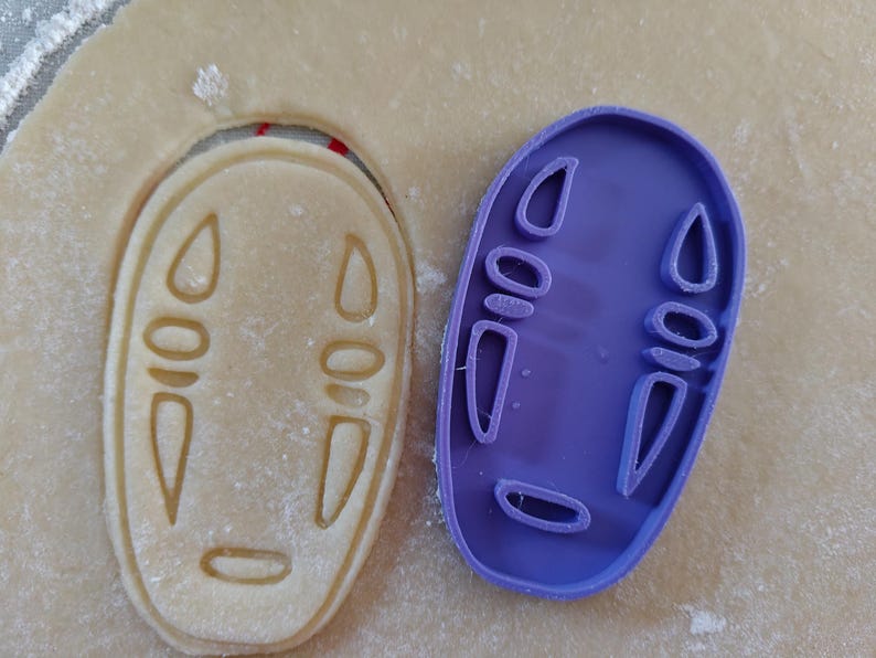 May include: A purple plastic cookie cutter in the shape of a character's face from the animated film, Spirited Away, next to a cookie cut out of the same shape. The cookie is light brown.