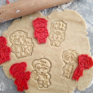 Puede incluir: Un conjunto de cortadores de galletas de plástico rojo con diseños de personajes de dibujos animados, junto con galletas cortadas. Un rodillo de madera descansa sobre la masa. Las galletas tienen forma de figuras de dibujos animados.