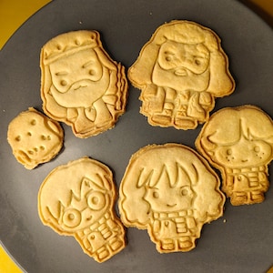 Peut inclure: Assortiment de biscuits cuits au four en forme de personnages d'une série fantastique populaire. Les biscuits sont dorés et disposés sur une assiette gris foncé. Les biscuits présentent des traits de visage et des vêtements détaillés.
