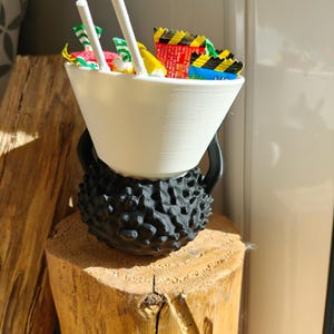 May include: A white, cone-shaped bowl filled with colorful candy wrappers and two white chopsticks, resting on a black, textured base. The base sits atop a wooden stump, creating a unique display.