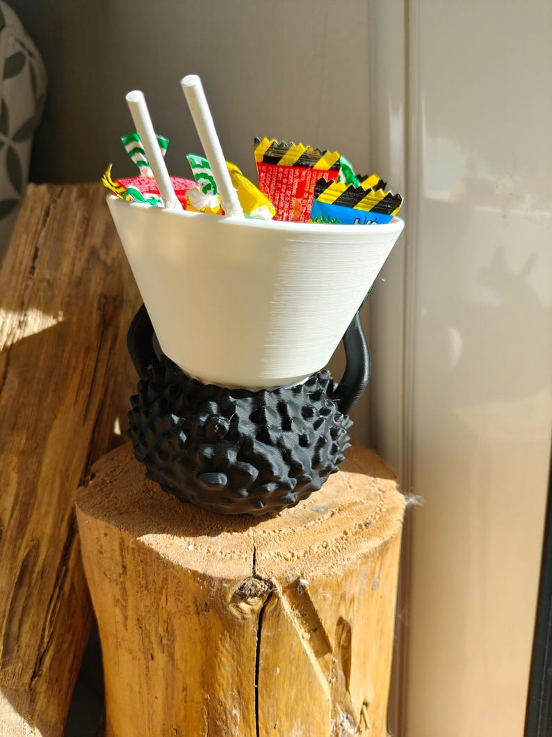 May include: A white, cone-shaped bowl filled with colorful candy wrappers and two white chopsticks, resting on a black, textured base. The base sits atop a wooden stump, creating a unique display.