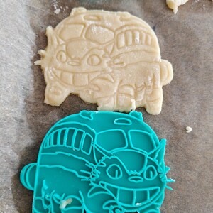 May include: A close-up shot of cookie cutters and cookies. The cookie cutters are teal and shaped like a bow and a bus with a cat face. The cookies are beige and shaped like a cat bus. The cookies are on parchment paper.