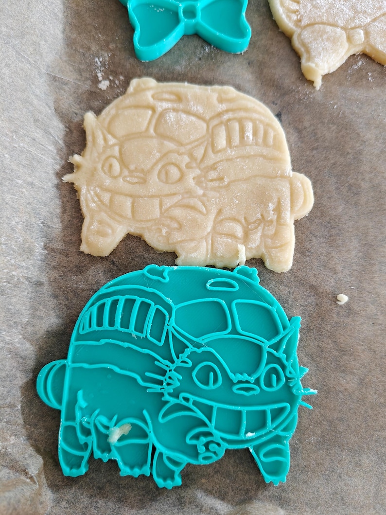 May include: A close-up shot of cookie cutters and cookies. The cookie cutters are teal and shaped like a bow and a bus with a cat face. The cookies are beige and shaped like a cat bus. The cookies are on parchment paper.