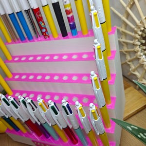 May include: A white tiered display stand with bright pink accents holds numerous multi-colored retractable ballpoint pens. The pens feature various barrel colors including yellow, blue, green, and patterned designs. A label reads Les Bics de Fabrina.