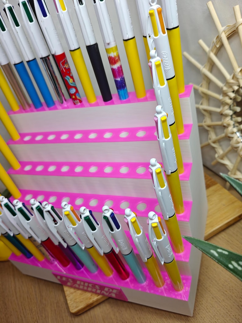 May include: A white tiered display stand with bright pink accents holds numerous multi-colored retractable ballpoint pens. The pens feature various barrel colors including yellow, blue, green, and patterned designs. A label reads Les Bics de Fabrina.
