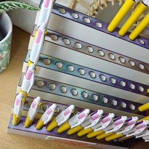 May include: A tiered display stand with an iridescent metallic finish featuring multiple circular cutouts for organizing pens. The stand holds several yellow and white multi-color ink pens with pink and purple accents arranged in the slots.