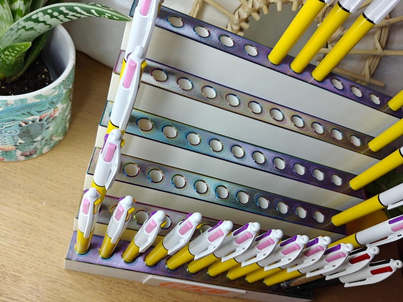 May include: A tiered display stand with an iridescent metallic finish featuring multiple circular cutouts for organizing pens. The stand holds several yellow and white multi-color ink pens with pink and purple accents arranged in the slots.