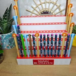 May include: A display of colorful pens with animal-themed toppers, arranged in a white and red tiered holder. The pens feature floral patterns and the holder has the text "LEGAMI DE VALENTIN". Decorative plants and a wooden fan are in the background.