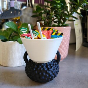 May include: A white bowl filled with colorful candies and two white sticks, resting on a black, textured base shaped like a kettlebell. The background includes green plants and kitchen utensils.