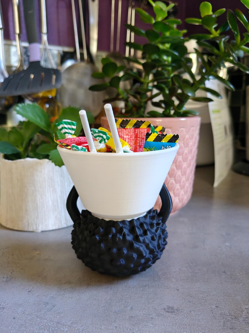 May include: A white bowl filled with colorful candies and two white sticks, resting on a black, textured base shaped like a kettlebell. The background includes green plants and kitchen utensils.