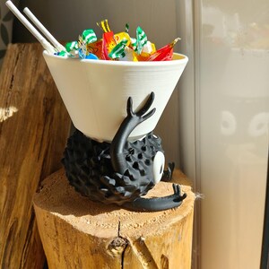 May include: A whimsical, black, spiky creature figurine with a white bowl-shaped head filled with colorful candies and two white chopsticks. The figurine is resting on a wooden stump, creating a playful and unique decorative piece.