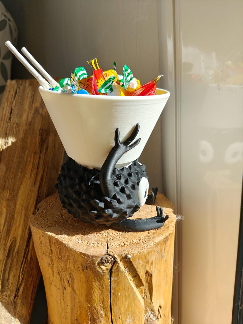 May include: A whimsical, black, spiky creature figurine with a white bowl-shaped head filled with colorful candies and two white chopsticks. The figurine is resting on a wooden stump, creating a playful and unique decorative piece.