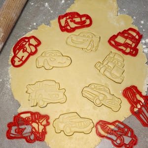 Puede incluir: Cortadores de galletas rojos con forma de coche sobre una masa de color claro, con formas de coche presionadas en la masa. Un rodillo de madera está en el fondo. Los cortadores de galletas tienen varios diseños de coches.