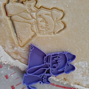 May include: A purple plastic cookie cutter in the shape of a cartoon witch with a cat, next to a cookie cut-out of a wizard. The witch cutter is on a gray surface with measurement markings. The cookie dough is light brown.
