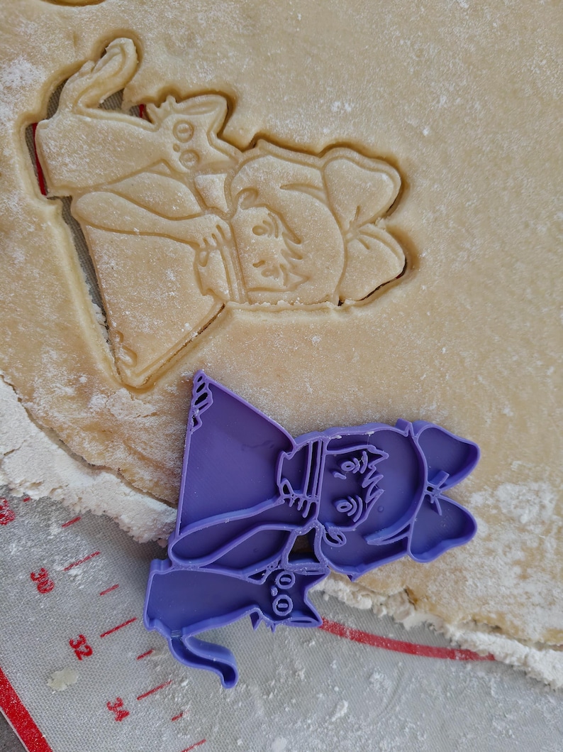 May include: A purple plastic cookie cutter in the shape of a cartoon witch with a cat, next to a cookie cut-out of a wizard. The witch cutter is on a gray surface with measurement markings. The cookie dough is light brown.